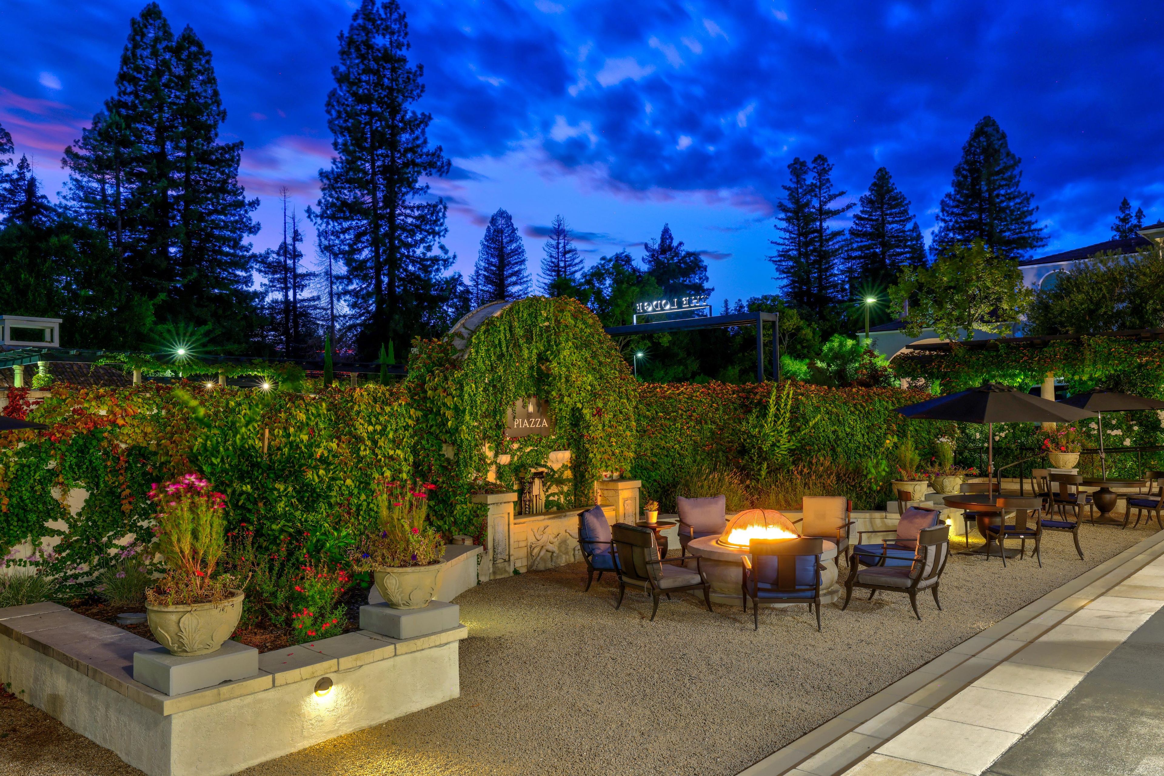 An outdoor patio area with seating and a fire feature, surrounded by lush greenery and trees.
