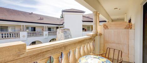 A balcony with a round table and wicker chair, overlooking a courtyard with buildings.
