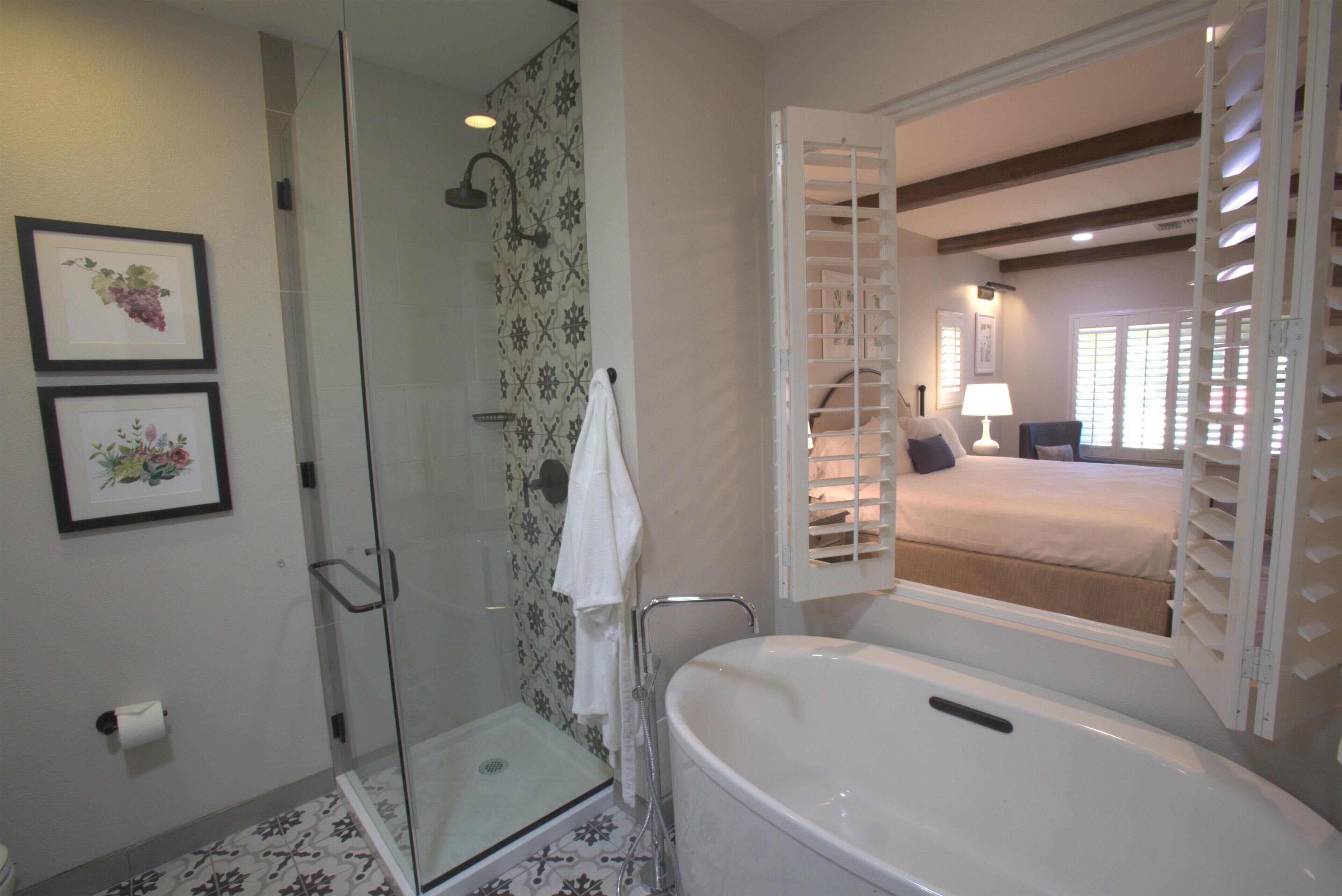 A bathroom with a glass shower enclosure, a bathtub, and a bed visible through an open door.