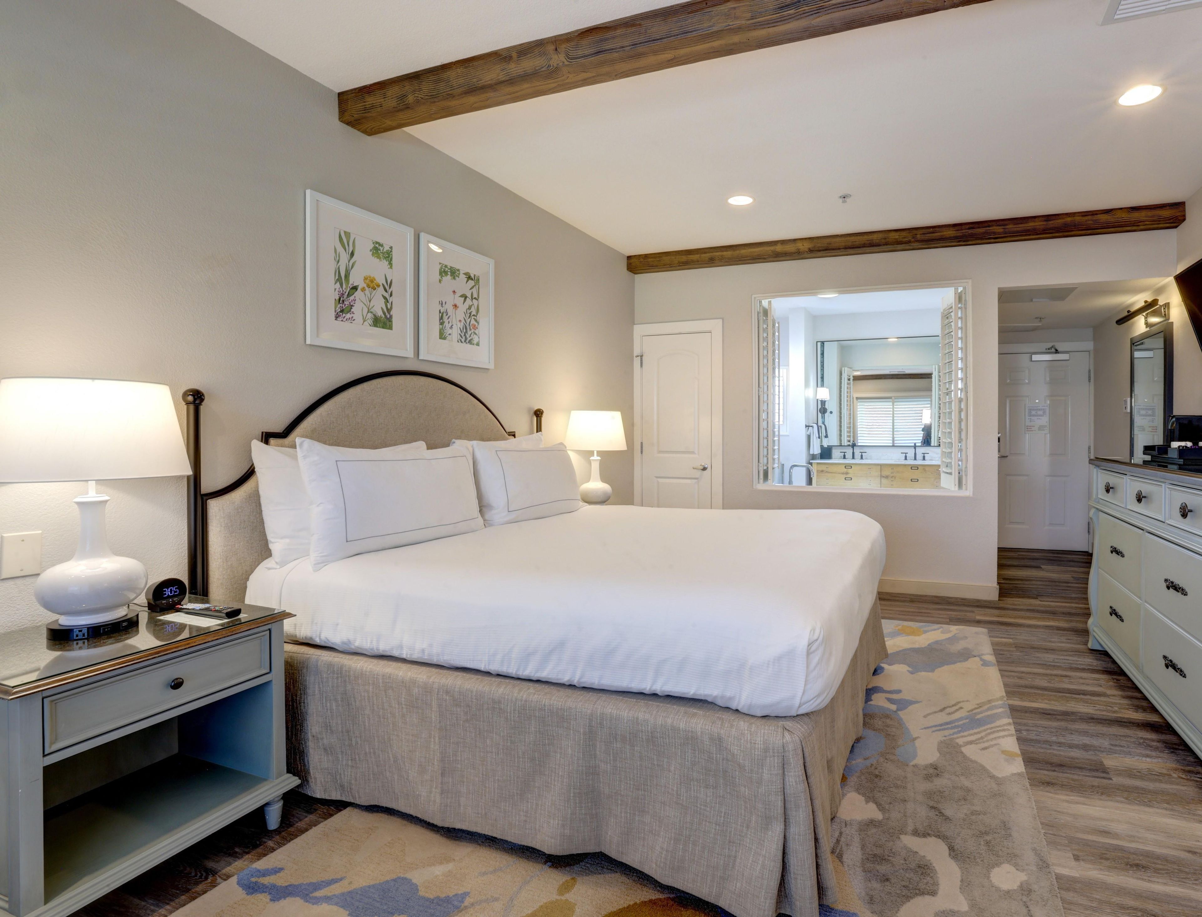 A bedroom with a bed, bedside tables, lamps, a nightstand, a mirror, and a door leading to a bathroom.