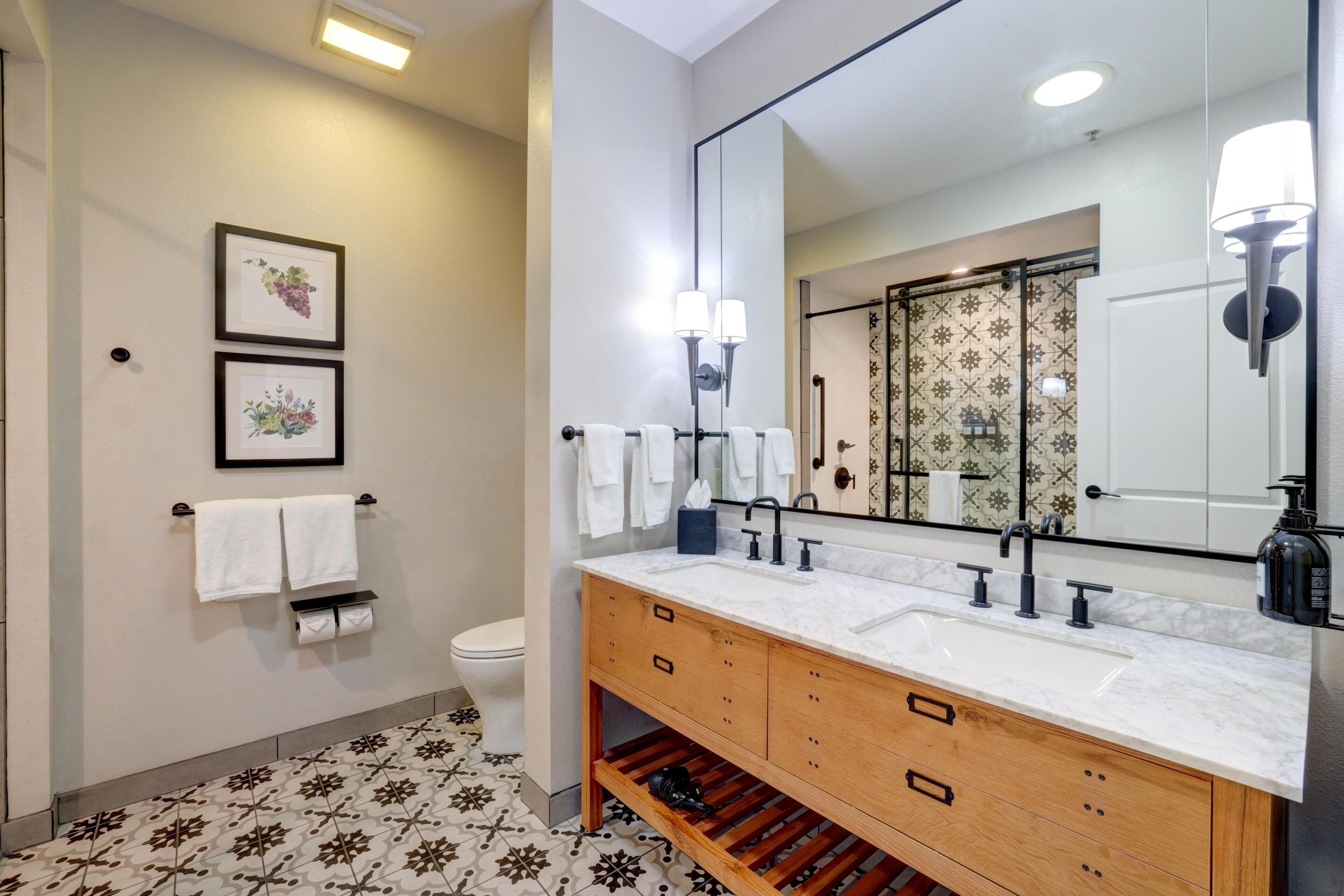 Un bagno con un grande specchio, lavabo bianco, mobile vanitario in legno, due quadri appesi al muro e piastrelle decorative sul pavimento.