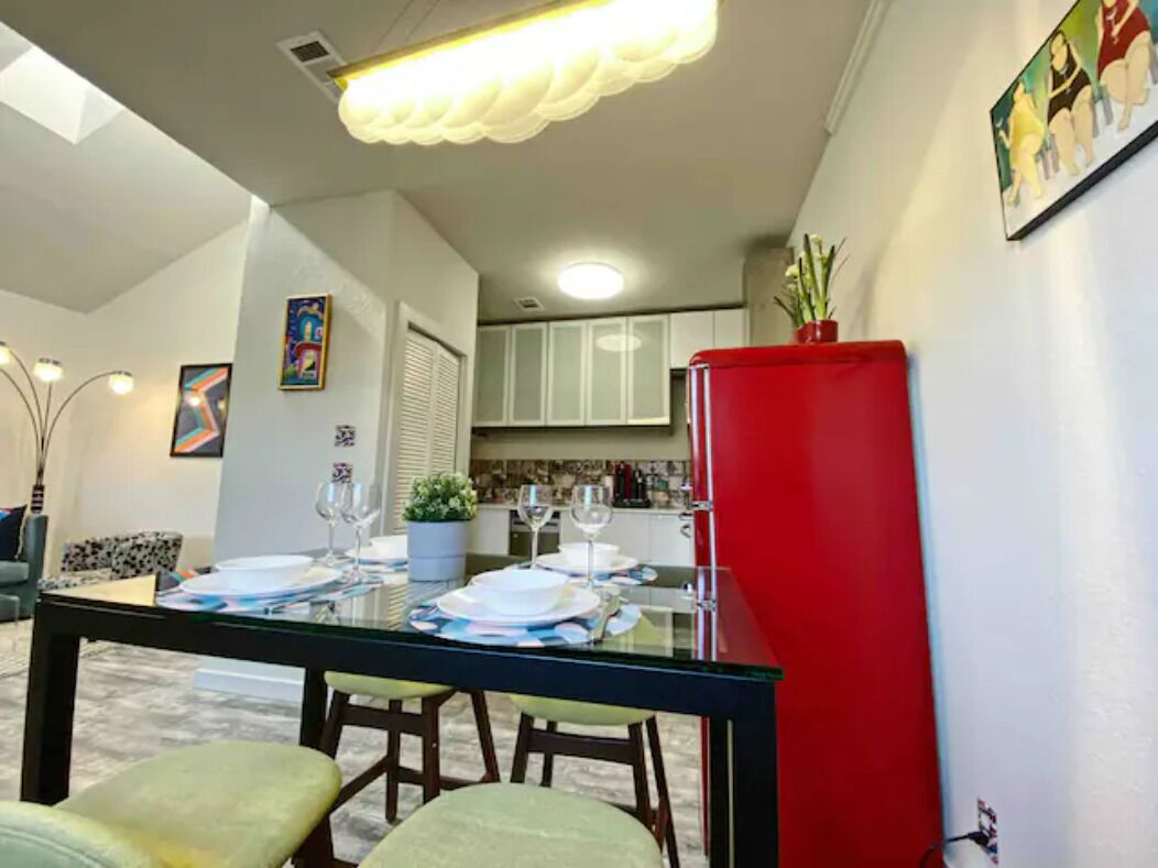 A dining area with a glass table, chairs, and a red refrigerator. There are framed artworks on the walls and a potted plant on the table.