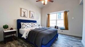 A bedroom with a bed, a nightstand, a ceiling fan, a window with curtains, and wall art.