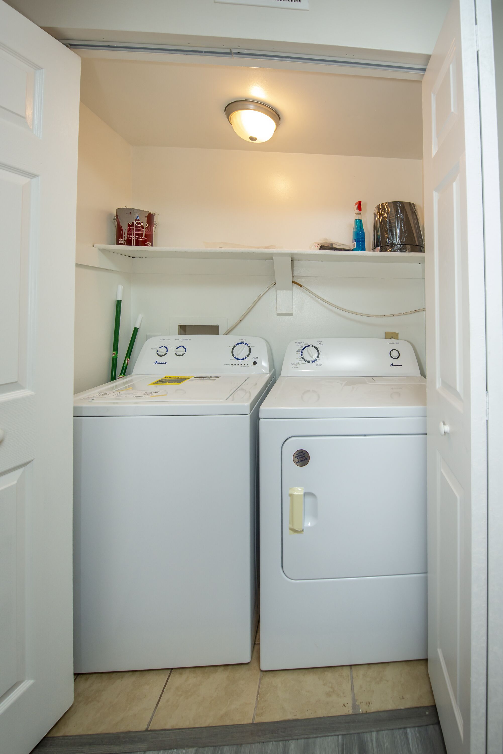 A laundry room with two white front-loading washing machines, a shelf with cleaning supplies, and a door leading to another room.