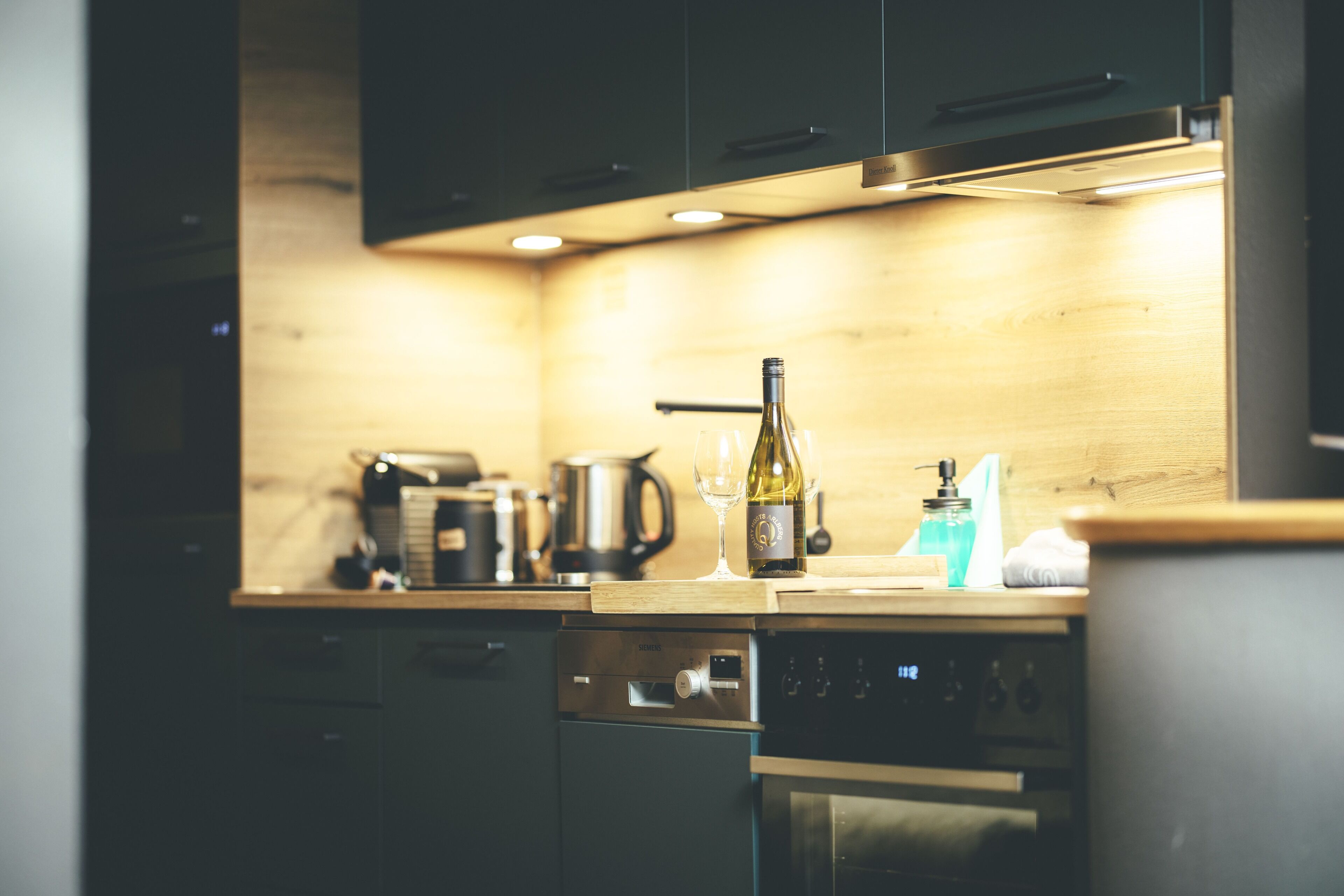 A modern kitchen with dark cabinets, a wooden countertop, a dishwasher, an oven, and a wine bottle.