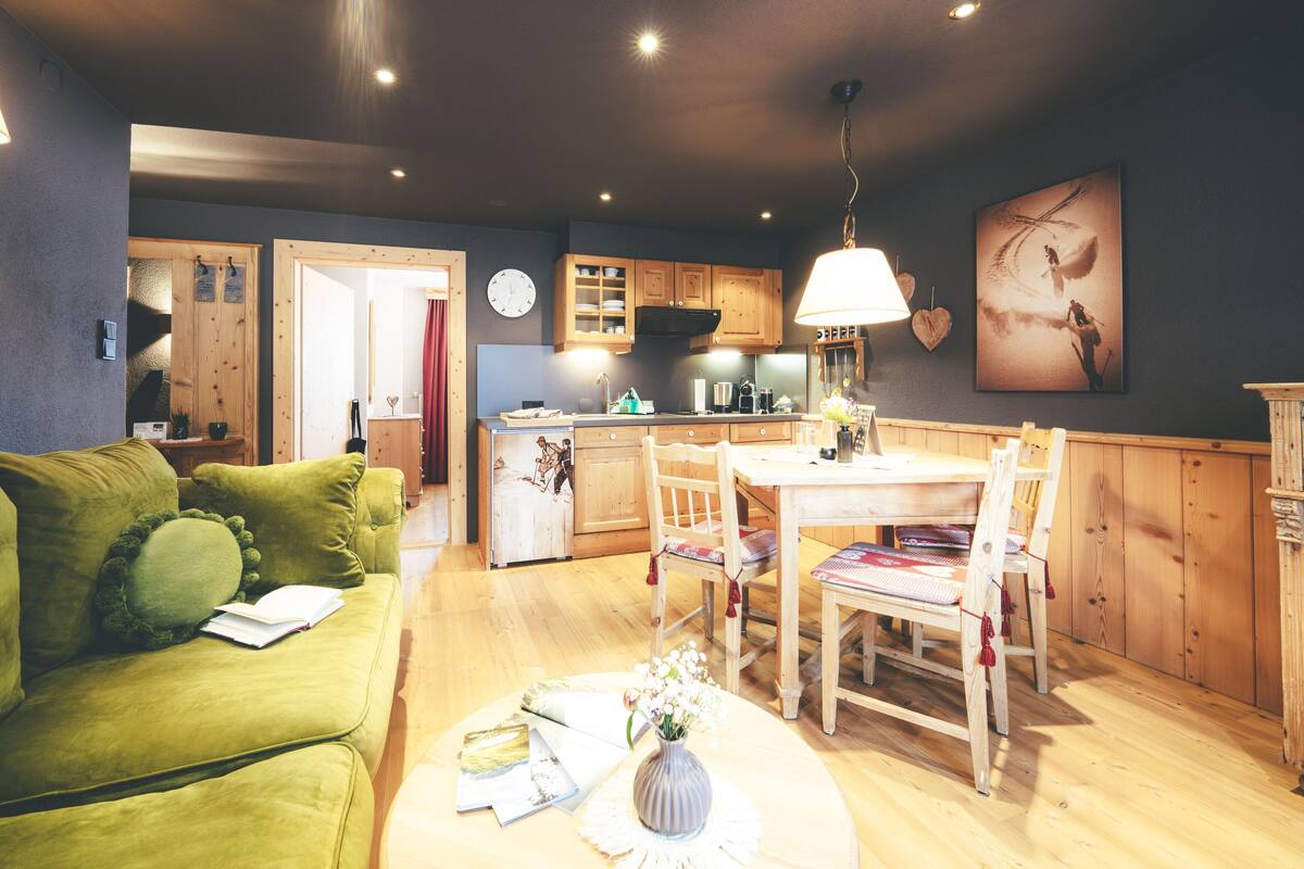A cozy living room with a green sofa, a dining table with chairs, and a kitchen area with wooden cabinets and a clock on the wall.
