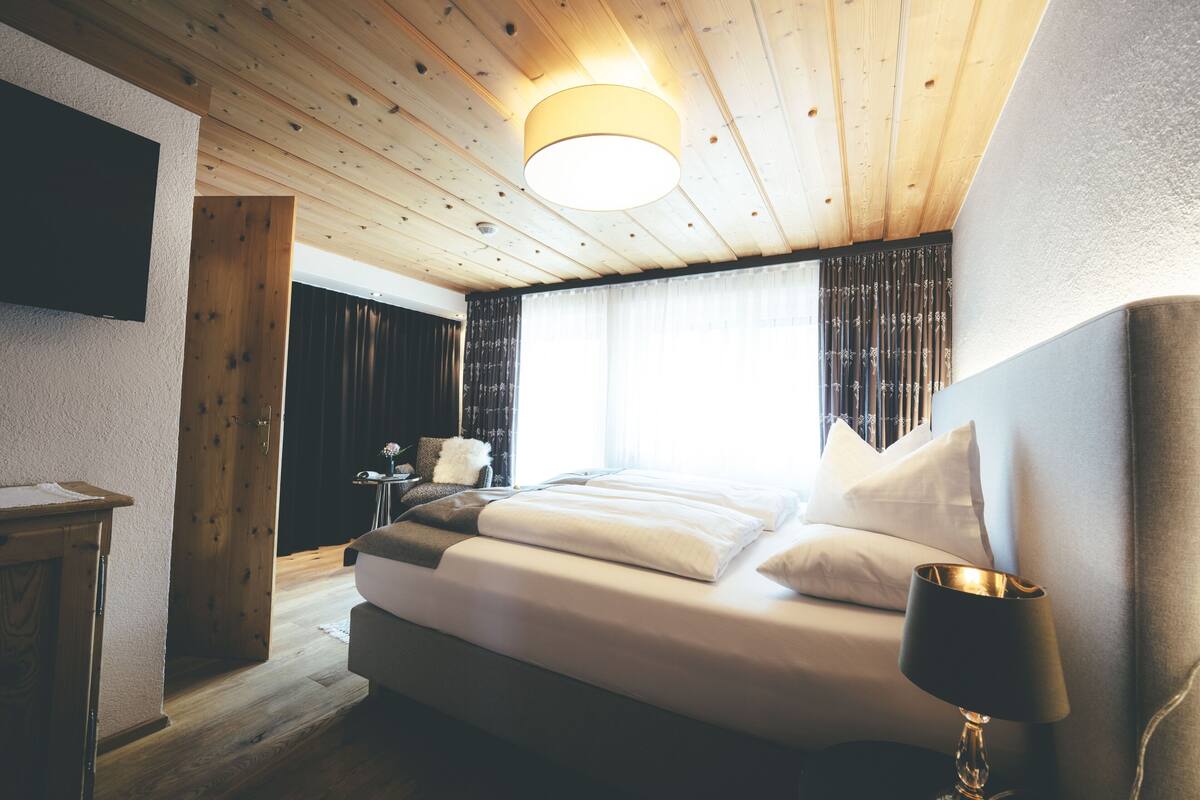 A modern hotel room with a wooden ceiling, a large bed with white linens, a bedside table with a lamp, and a TV mounted on the wall.
