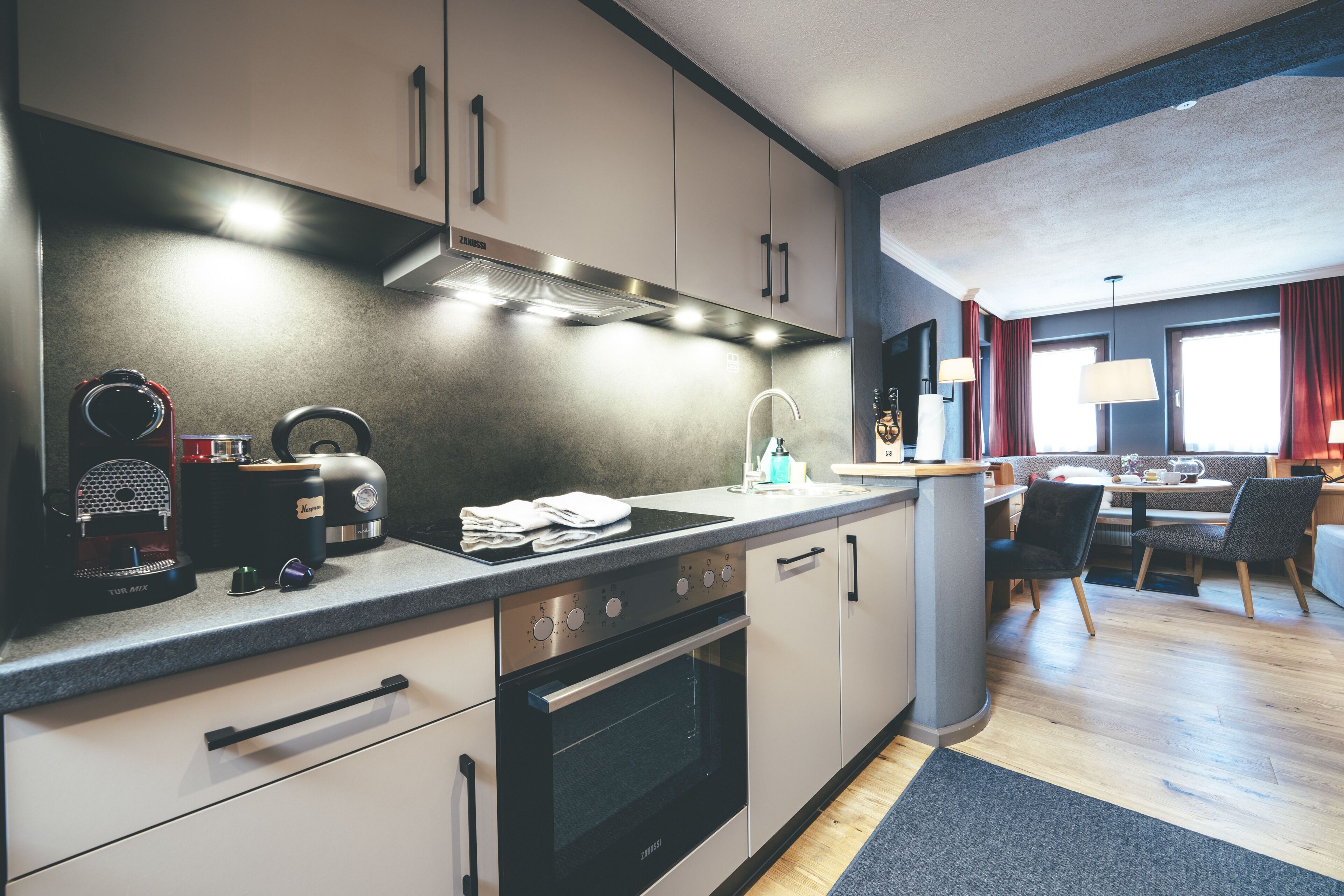 A modern kitchen with a built-in oven, a coffee maker, and a sink. There is a dining area with a table and chairs.