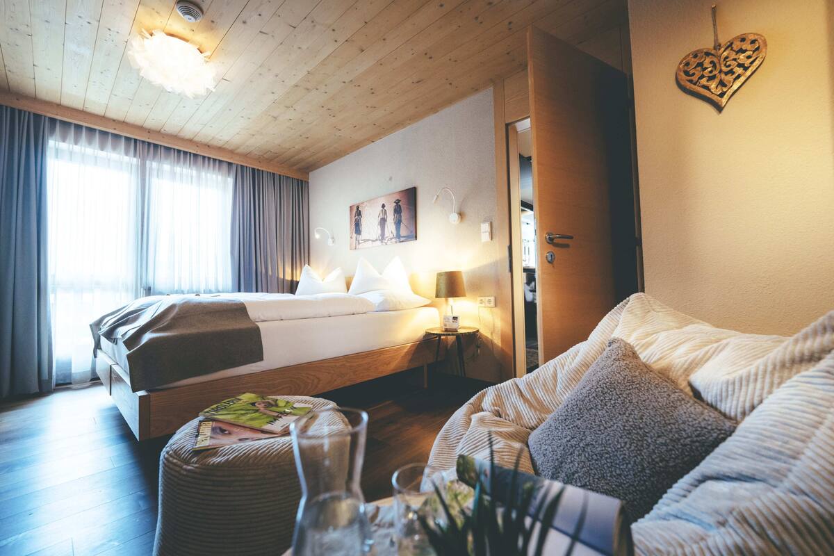 A hotel room with a wooden ceiling, a bed with a white comforter, a sofa with cushions, a small table with a vase and magazines, and a painting on the wall.