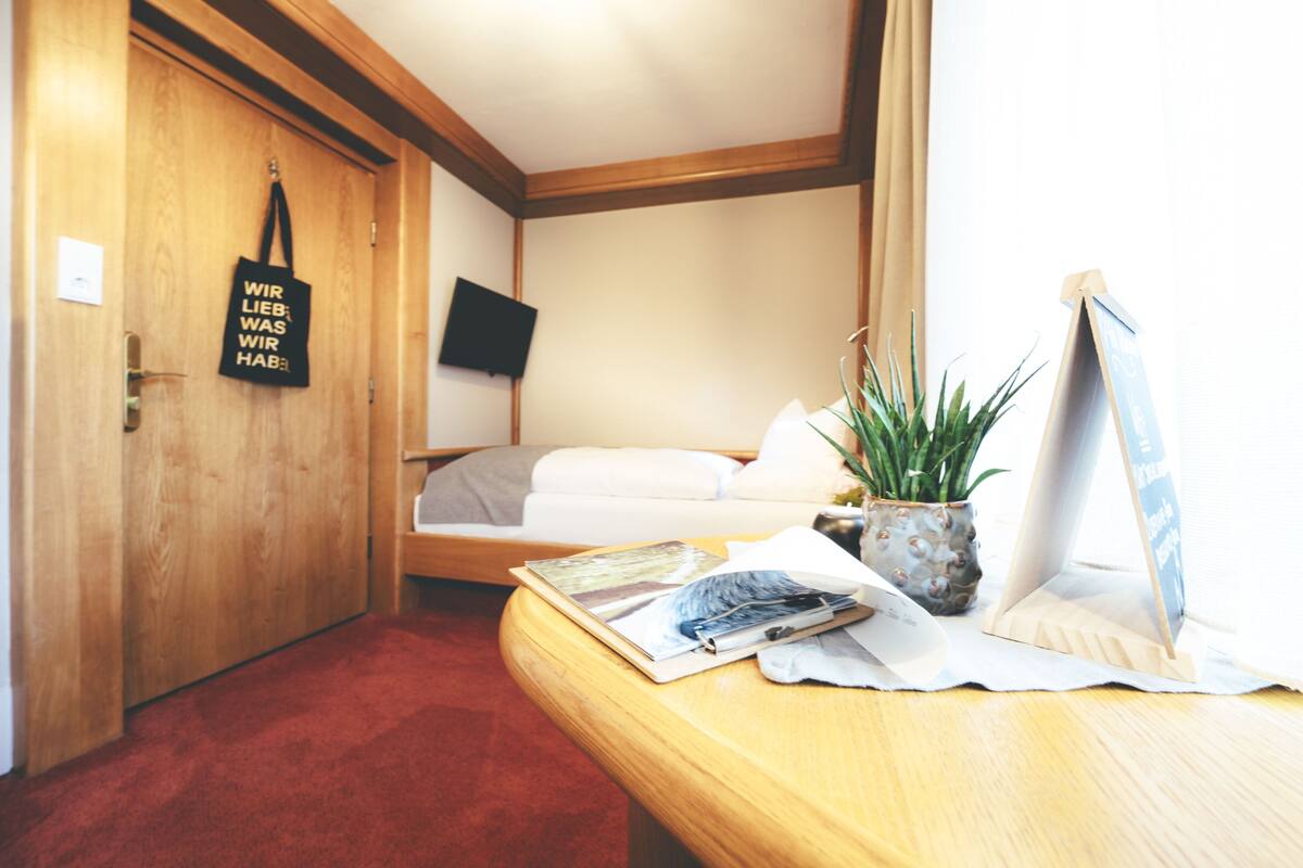 A hotel room with a wooden door, a bed with white bedding, a small table with a plant and a sign, and a TV mounted on the wall.