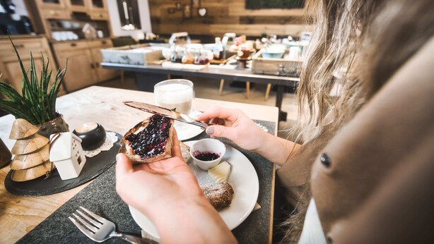 A person holding a slice of bread with jam, a small bowl of jam, and a plate with a pastry. - Lärchenhof - Premium Apartments & Zimmer (Sankt Anton am Arlberg)
