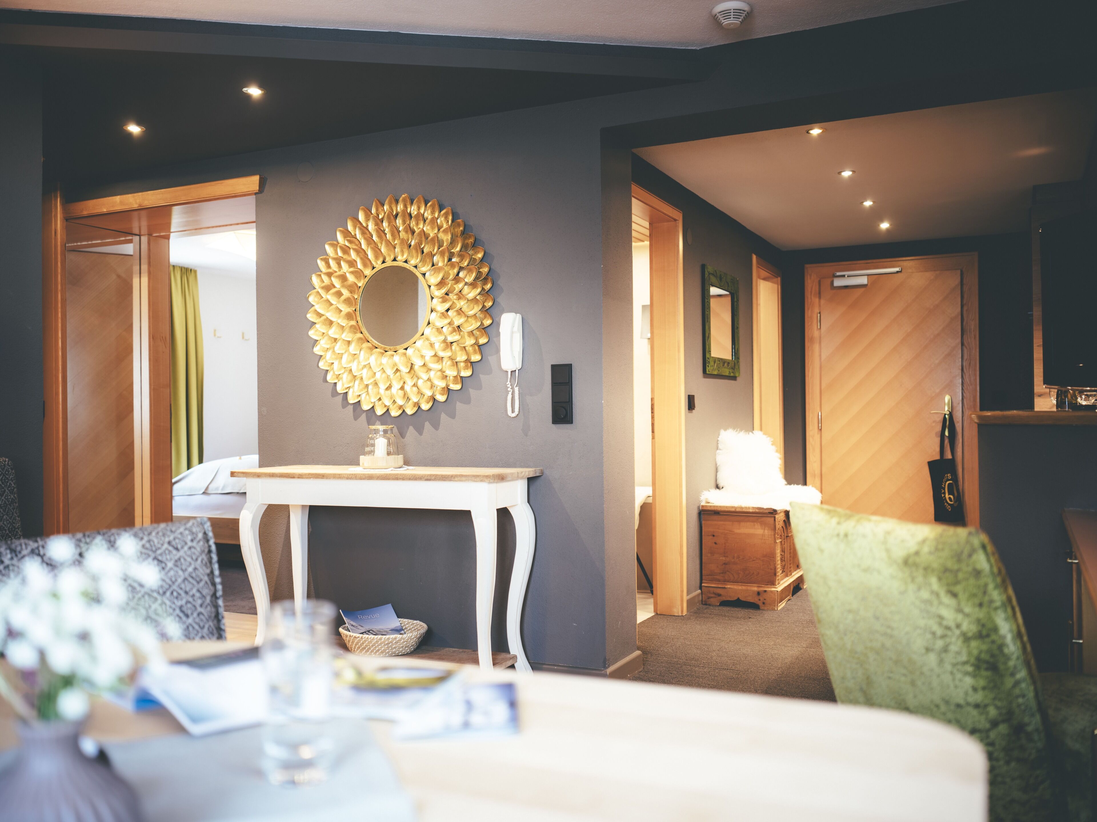 A hotel room with a round mirror, a bedside table, a chair, and a wooden wardrobe.