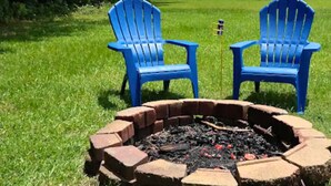 Un hoyo de fuego circular de ladrillo con dos sillas Adirondack azules orientadas hacia él.