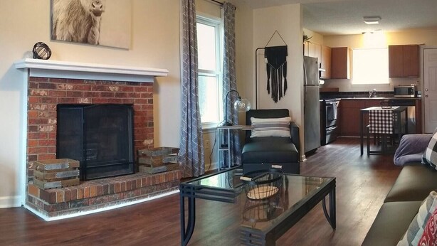 Una sala de estar con chimenea, una mesa de centro de vidrio, un sofá y una cocina al fondo.