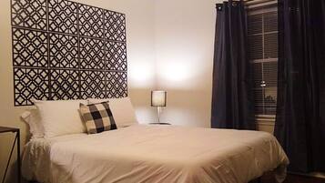 A neatly made bed with white bedding and a checkered pillow, a wall-mounted decorative panel, a bedside lamp, and a window with dark curtains.