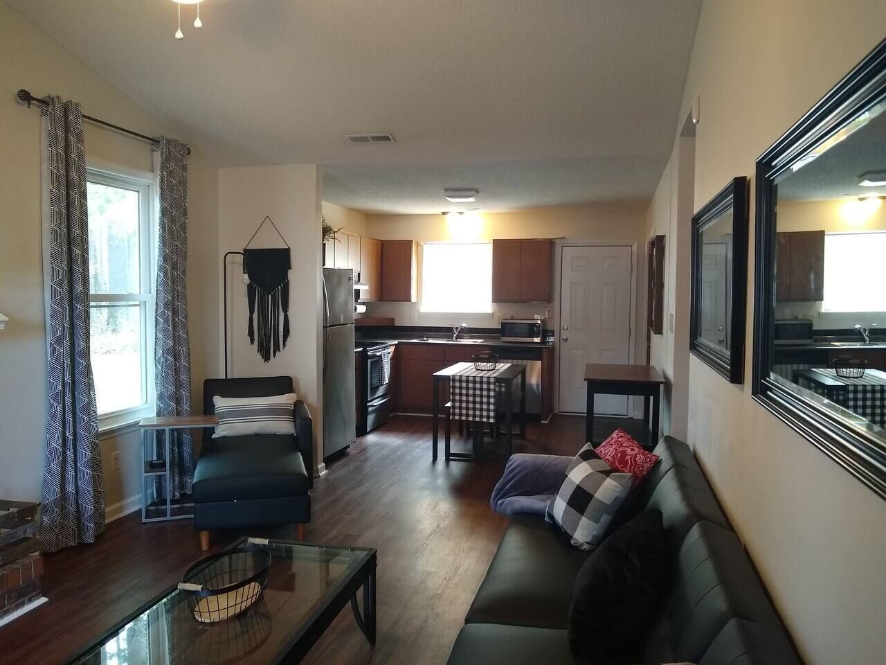 A living room with a sofa, a glass coffee table, and a dining area with a table and chairs. There is a kitchen in the background with a microwave and a refrigerator.