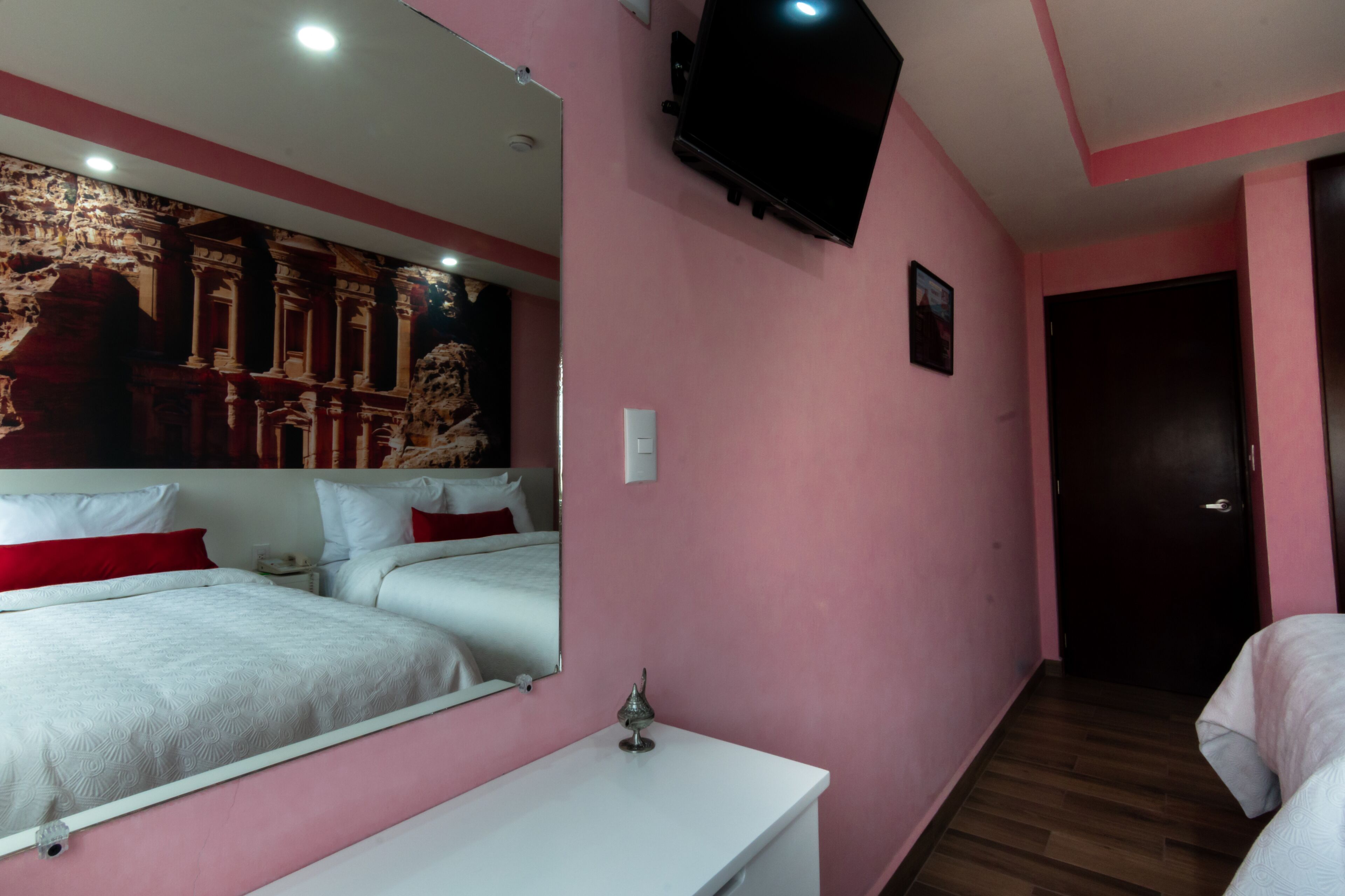A hotel room with pink walls, a large bed with white bedding, a bedside table, a TV mounted on the wall, and a door leading to another room.