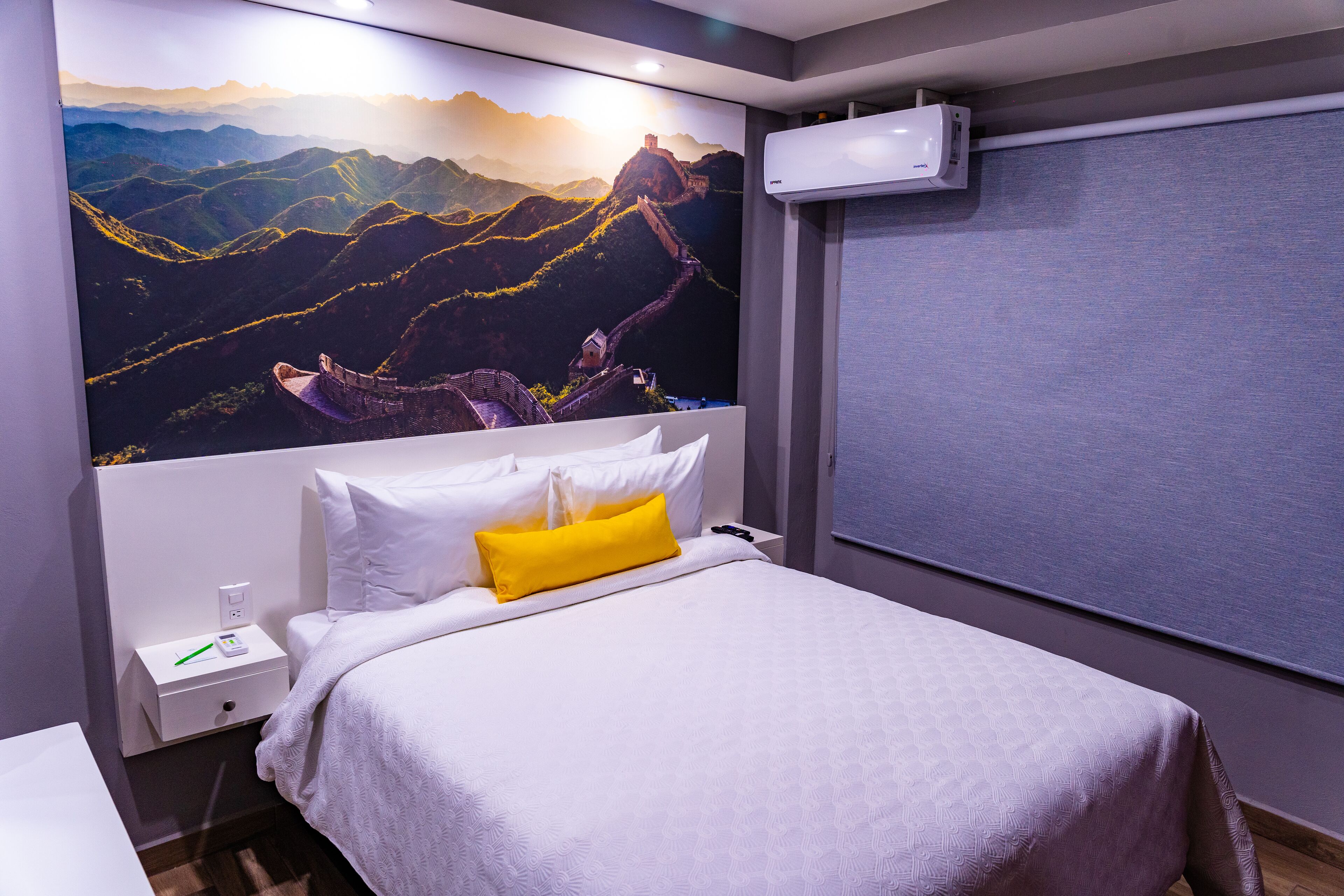 A hotel room with a large wall mural of the Great Wall of China, a bed with white and yellow bedding, a bedside table with a phone and a small item, and a window with a blind.
