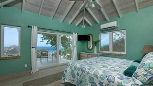 A bedroom with a large bed, a ceiling fan, and a view of the sea through a sliding glass door. - Villa Colada, a 4-Bedroom St. John Villa Above Rendezvous Bay (St. John)