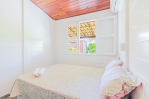 Une petite chambre avec un lit simple, un plafond en bois, une fenêtre donnant sur la verdure et un lit soigneusement fait avec une couvre-lit à motifs floraux.