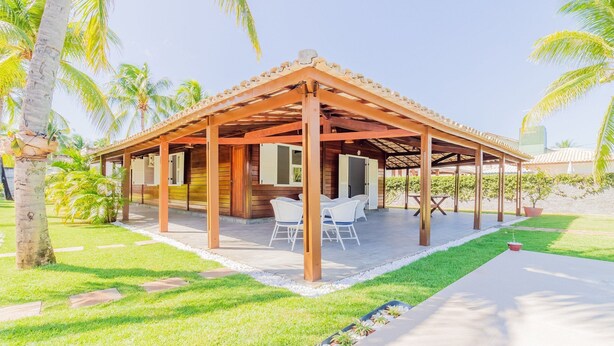 A covered outdoor area with a wooden structure, a table with chairs, and a small garden area with palm trees. - GB69 Excellent 4/4 house 400m from the beach (Camaçari)