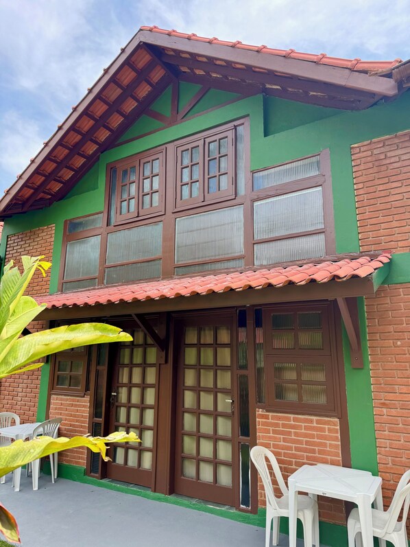 A two-story house with a green facade, brown windows, and a tiled roof. There are white plastic chairs and a table outside. - Chales da Lua Juquehy (São Sebastião)