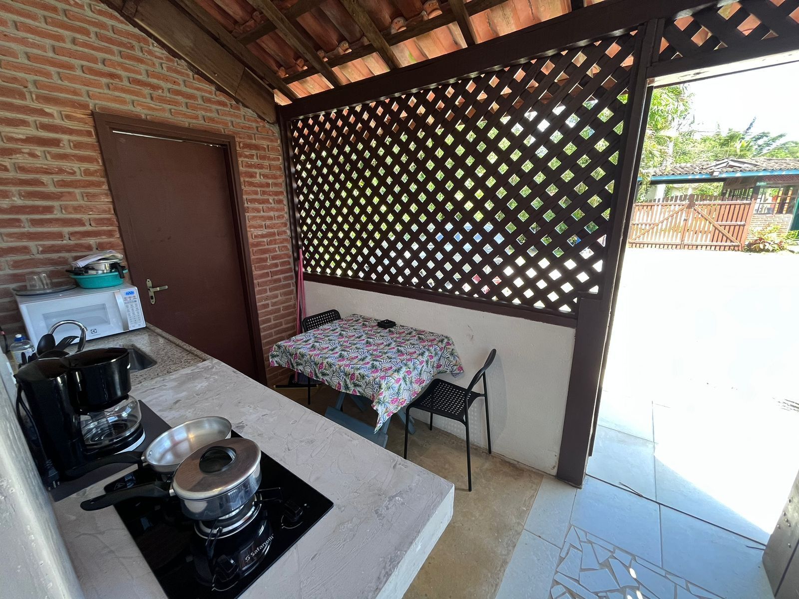 A compact kitchen with a gas stove, a microwave, and a sink. There is a table with a patterned tablecloth and a chair. A wooden lattice partition separates the kitchen from the outdoor area.