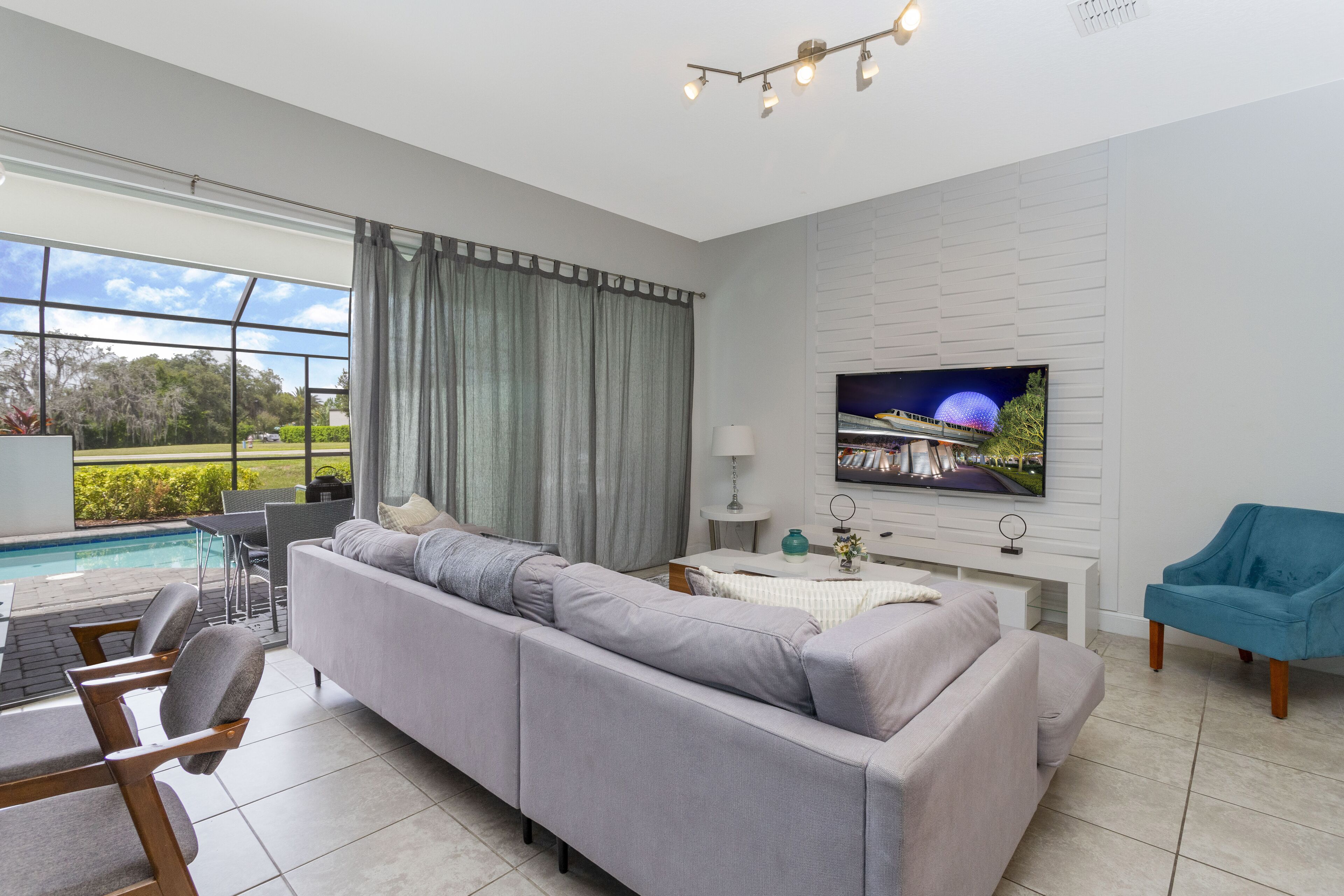 Una sala de estar moderna con un sofá gris, un sillón azul, un televisor montado en una pared blanca y una piscina visible a través de ventanas amplias.