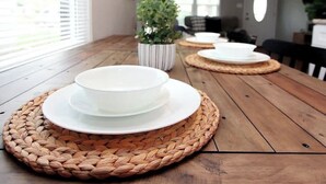 Une table à manger avec de l’assiette blanche et des sets de table en fibres tressées, une plante en pot, et une vue sur un espace de vie avec un canapé et une porte au fond.