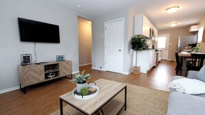 Un salon avec un téléviseur, une table basse en bois et un canapé. Au fond, on aperçoit un coin cuisine avec un four à micro-ondes et un évier.