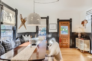 Un comedor con una larga mesa de madera, cojines blancos y negros a cuadros, una lámpara colgante tejida y un perro blanco.