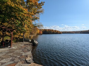 A stone pier with a gazebo and seating area overlooking a calm lake, surrounded by trees with autumn foliage.