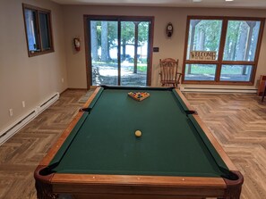 A pool table with a cue ball, a wooden chair, and a welcome sign by a large window.