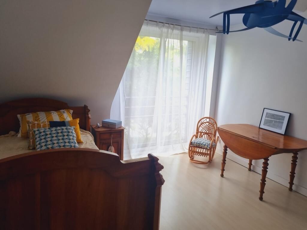 Ein Schlafzimmer mit einem Bett, einem Nachttisch aus Holz, einem Korbstuhl, einem Holztisch und einem Fenster mit transparenten Vorhängen.