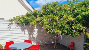 Eine Terrasse mit Tisch und roten Stühlen, einer weißen Wand und einem Baum mit lila Blüten.
