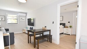 A modern living room with a wooden dining table, a grey sofa, and a TV mounted on the wall.