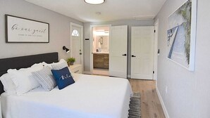 A bedroom with a bed, a bedside lamp, a framed quote on the wall, and a door leading to a bathroom.
