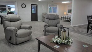 A room with two grey recliners, a wooden coffee table with a floral centerpiece, and a view of a bedroom with a checkered bedspread. - Country View Lodging In the heart of Amish Country (Baltic)