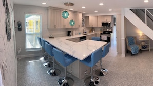 A modern kitchen with a central island, a dining area with blue chairs, a staircase, and a living room with a brown sofa and blue cushions. - Neptune's Grotto Waterfront Retreat (Spring Hill)