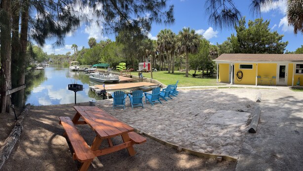 A lakeside picnic area with a wooden table and chairs, a small yellow building, and boats docked nearby. - Neptune's Grotto Waterfront Retreat (Spring Hill)