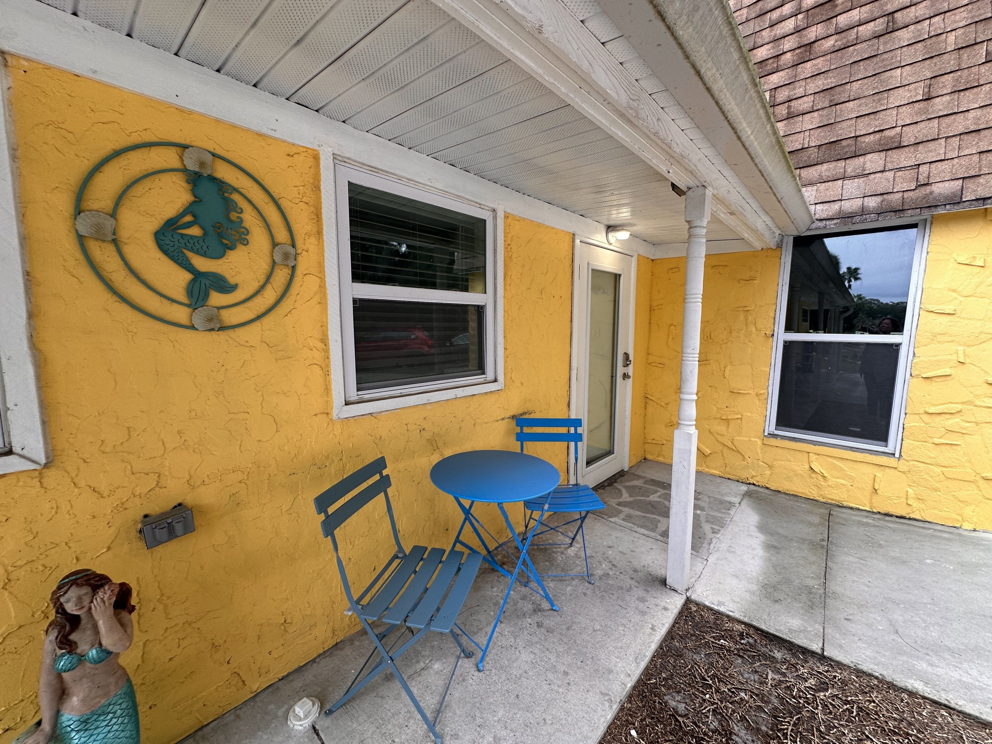 A yellow exterior wall with a mermaid sculpture, a window with blinds, and a blue folding chair.