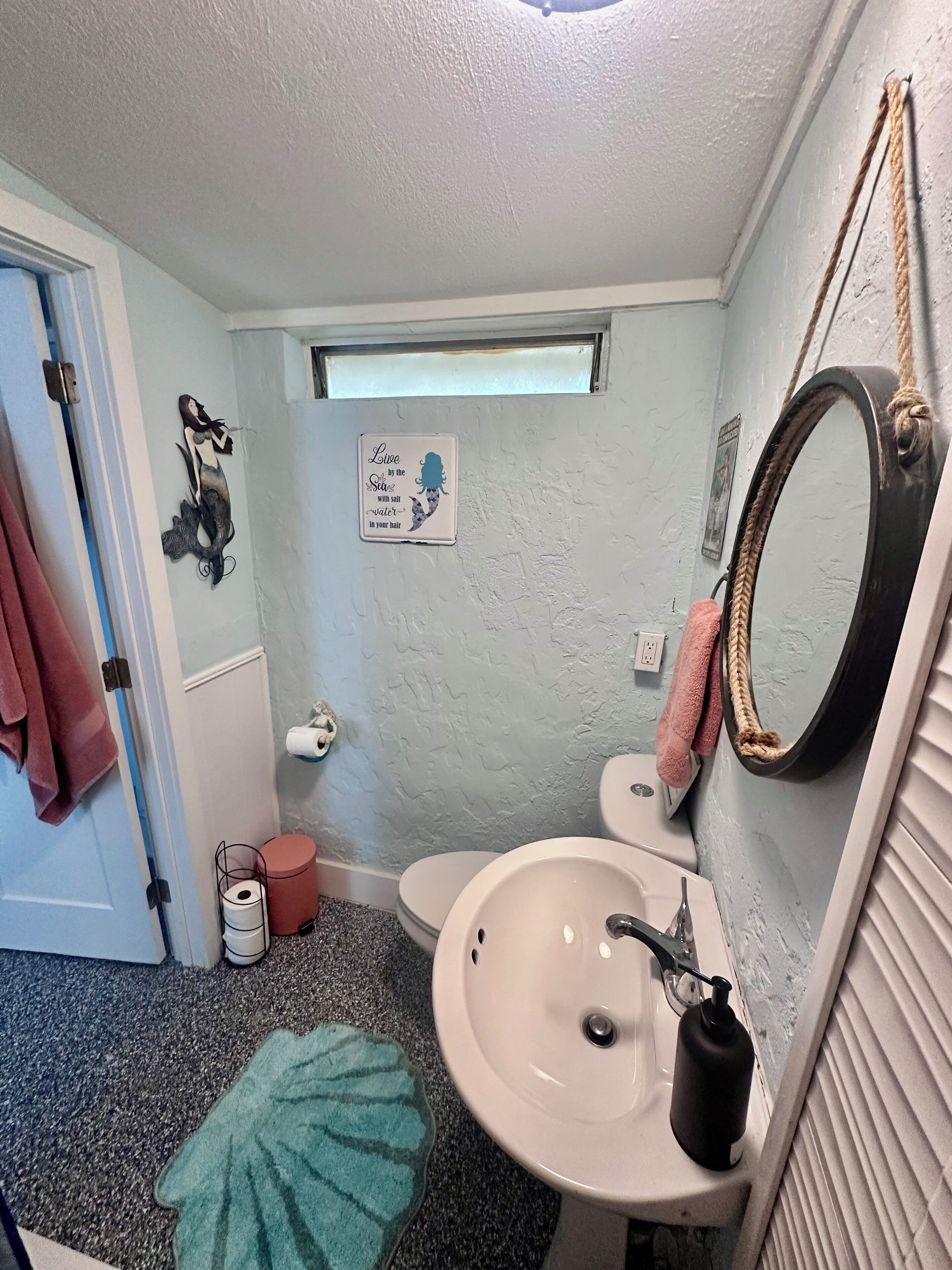 A bathroom with a sink, toilet, and a small window. There is a towel rack with towels, a mirror, and a decorative sign on the wall.