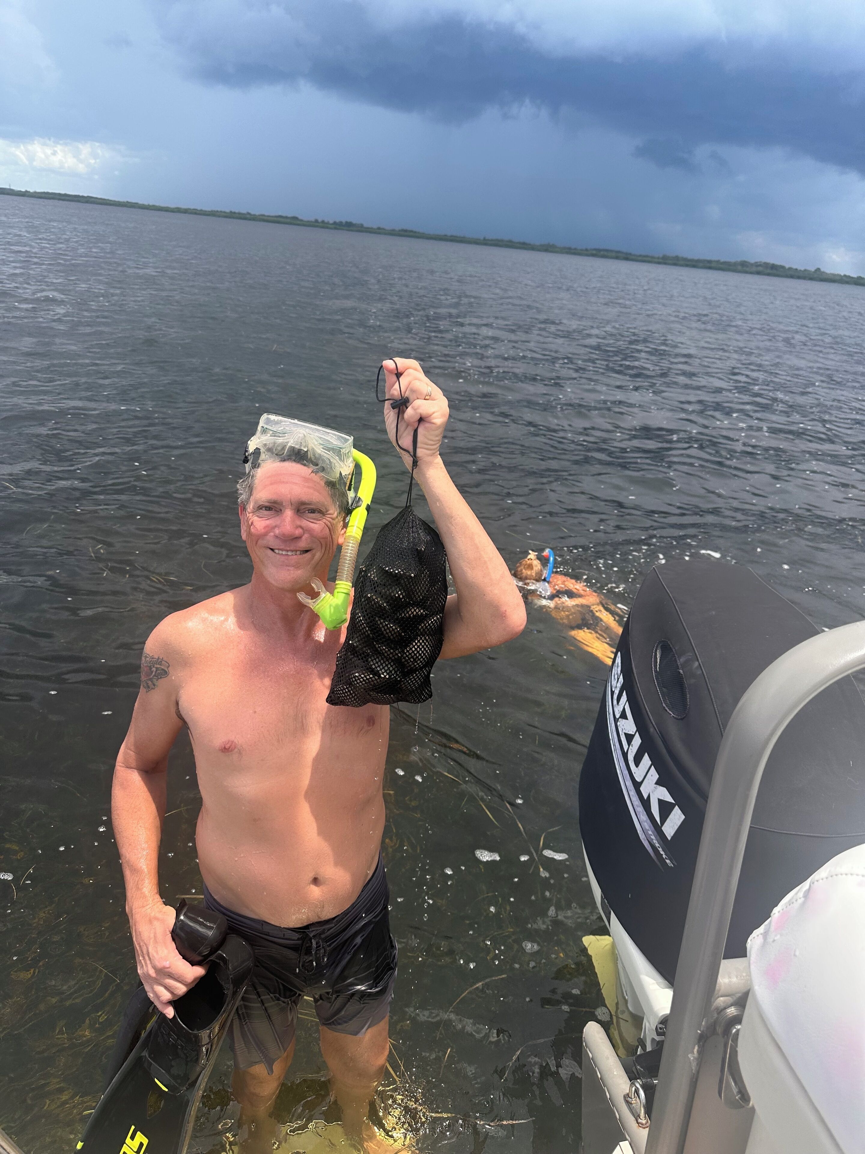A person holding a black mesh bag and a green snorkel, standing in shallow water near a boat with a Suzuki logo.