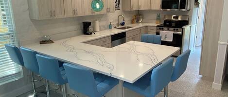 A modern kitchen with a marble island, blue bar stools, stainless steel appliances, and wooden cabinets.