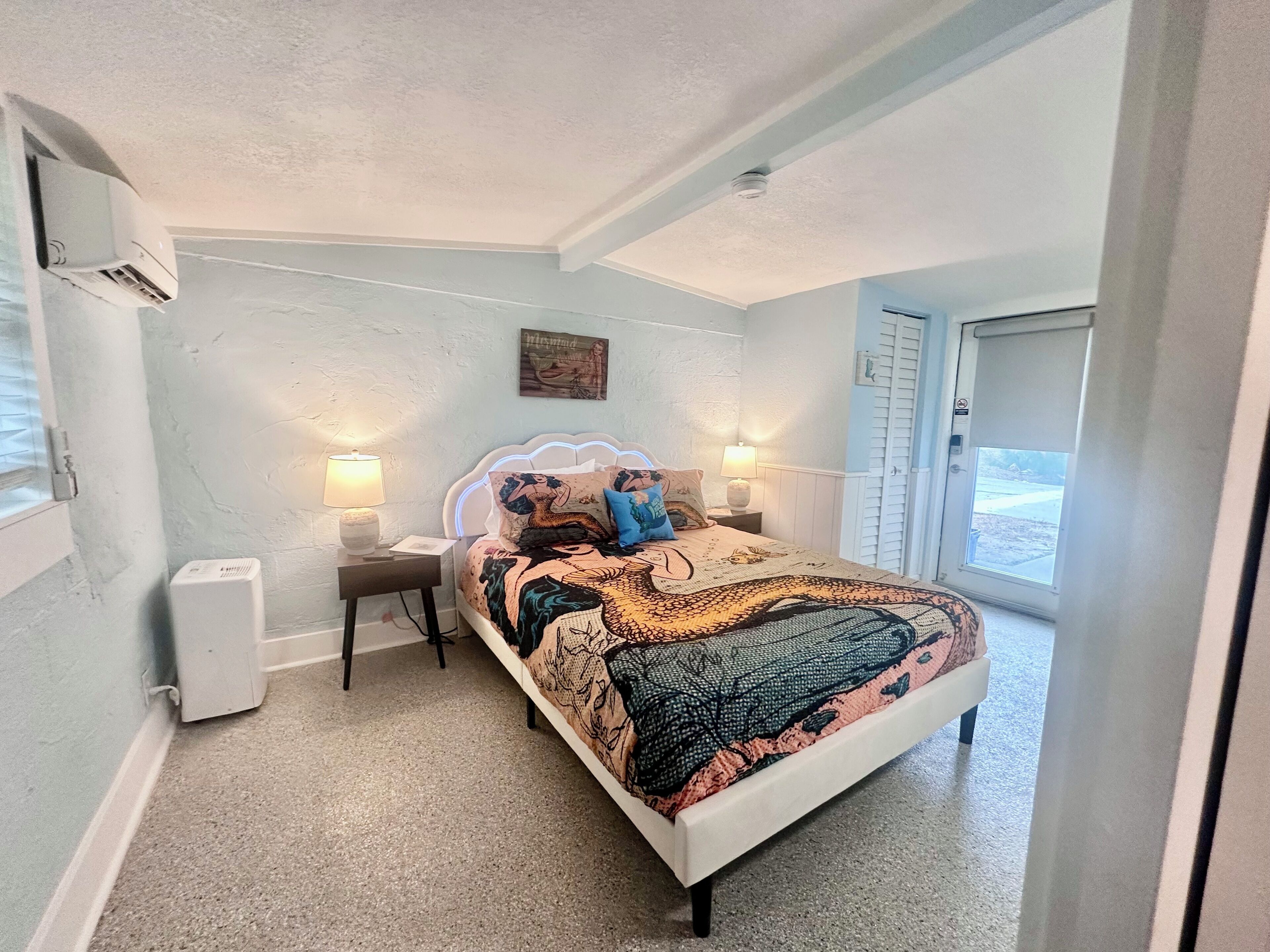 A bedroom with a bed, two bedside lamps, a wall-mounted air conditioner, a door, and a window.