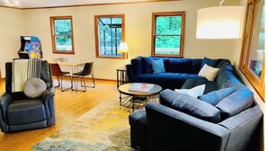 A living room with a sectional sofa, a coffee table, a rug, and a TV. There are windows with views of greenery. - SkyBarn on Rocky Knob - Views, Relaxation, Adventure, Game Room & Hot Tub (Zionville)