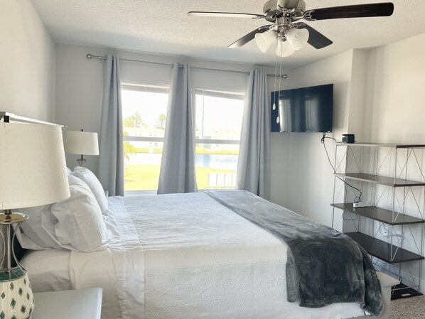 A hotel room with a bed, a ceiling fan, a TV mounted on the wall, and a view of a green landscape through the window. - Large , CLEAN townhome with pool view, next to Sports Complex (Panama City Beach)