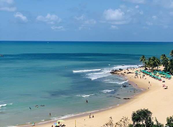 A beach with clear blue water, people swimming and sunbathing, and palm trees.
