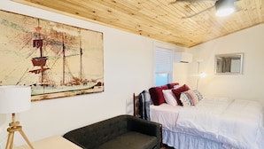A room with a wooden ceiling, a bed with white bedding, a dark sofa, a small table with a lamp, and a painting of a ship on the wall. - Breathtaking Views!  (Matlacha)