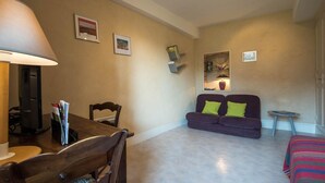 Una sala de estar con una mesa de comedor de madera, un sofá púrpura con cojines verdes, una pequeña mesa redonda y una lámpara.